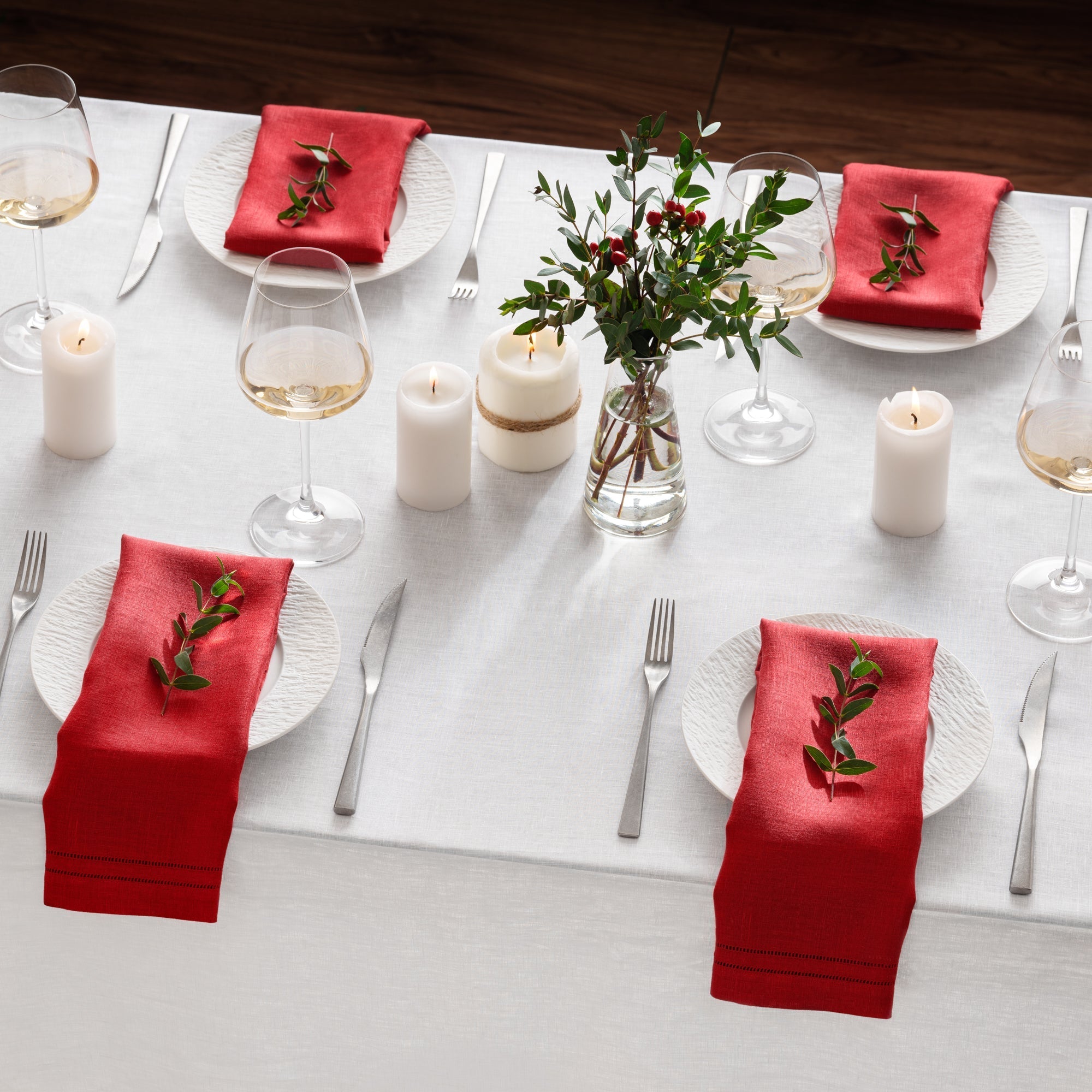 red linen napkins with elegant double hemstitching lines setting on winter christmas tablescape for four with beautiful white linen table cloth, ceramic plates and silver cutlery set, candles, fresh flowers and wine glass
