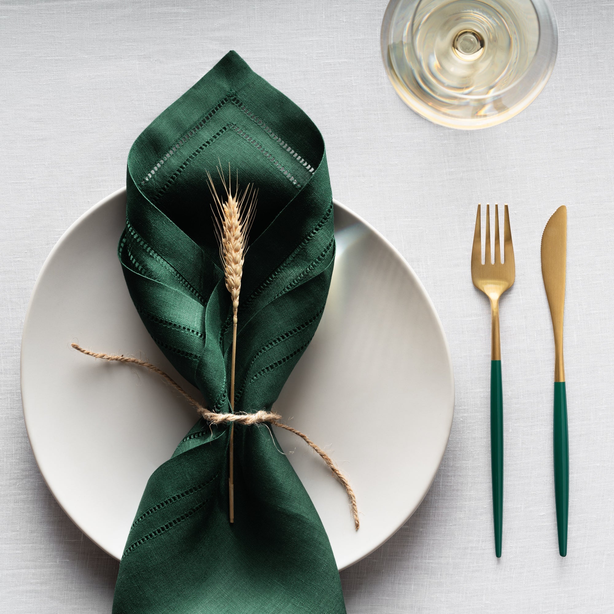 linen double hemstitched napkin in forest green setting on white linen table cloth and matching green and gold cutlery