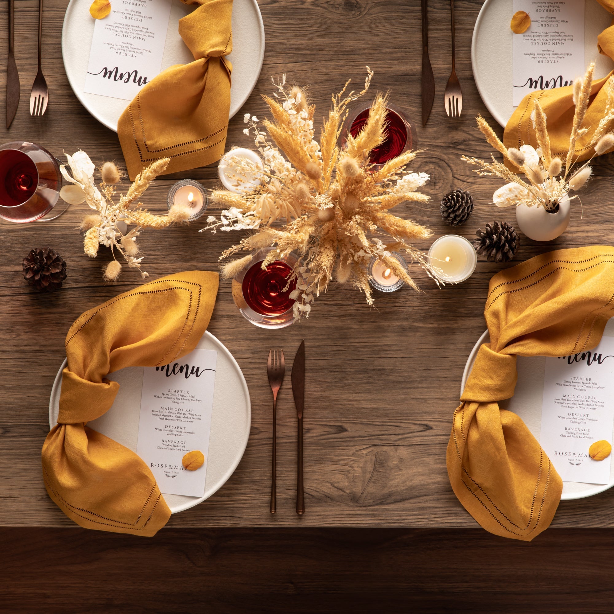 yellow linen napkins with elegant double hemstitching lines setting on beautiful thanksgiving tablescape for four on farmhouse table with ceramic plates and gold cutlery set, twilight candles, dried flowers and wine glass